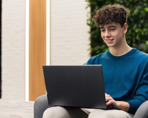 A young smiling man working using a laptop while sitting in a chair in an office
